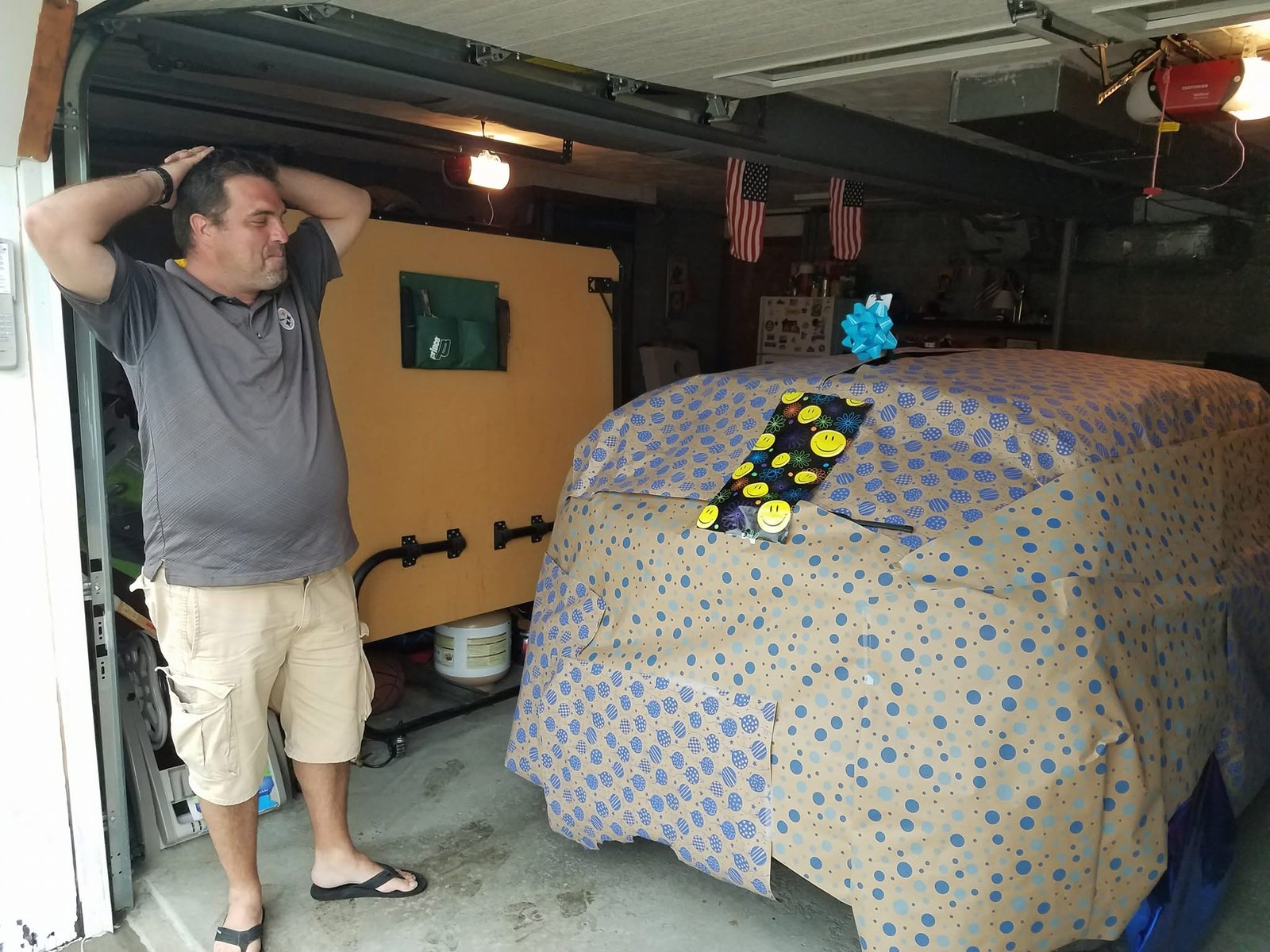 Tom in a garage with both hands on his head, staring at a car wrapped in wrapping paper.