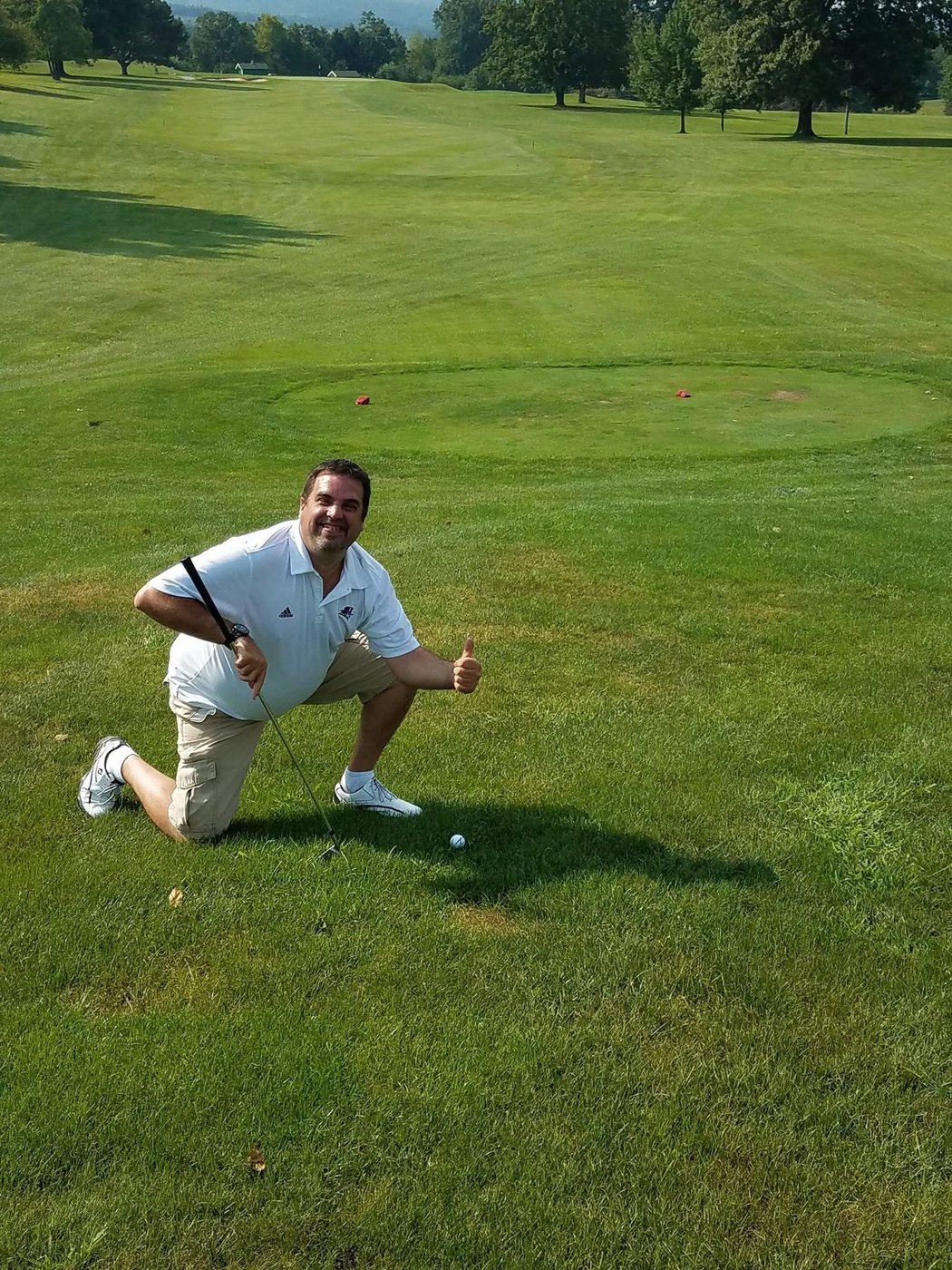 Tom kneeling on a golf course giving a thumbs up next to a ball in the rough.